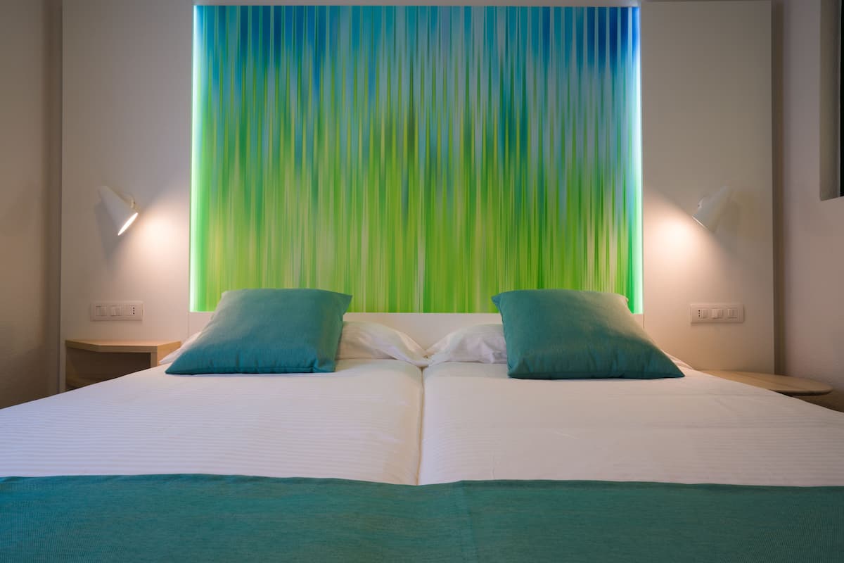 A double bed with white linens and two teal pillows, a green-striped curtain, and two wall-mounted lamps.
