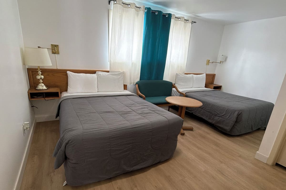 A hotel room with two beds, a chair, a small table, and a lamp.