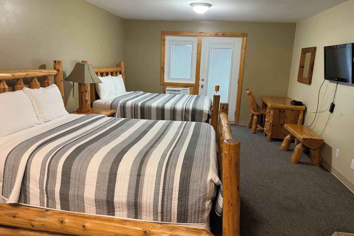 A room with two beds, a wooden headboard, a desk, a chair, a television, and a door with windows.