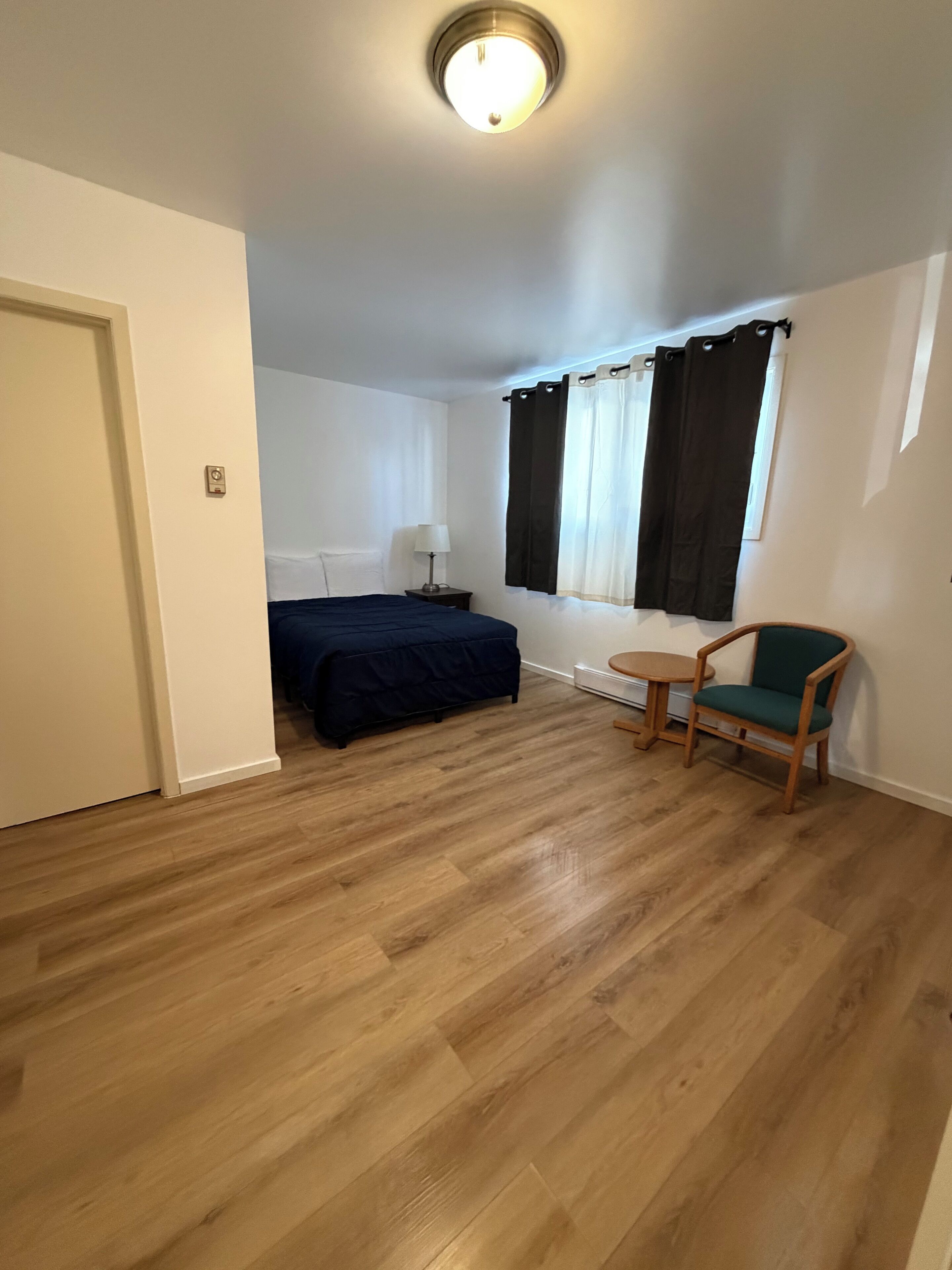 A room with a bed, a chair, a small table, a lamp, and a window with curtains.