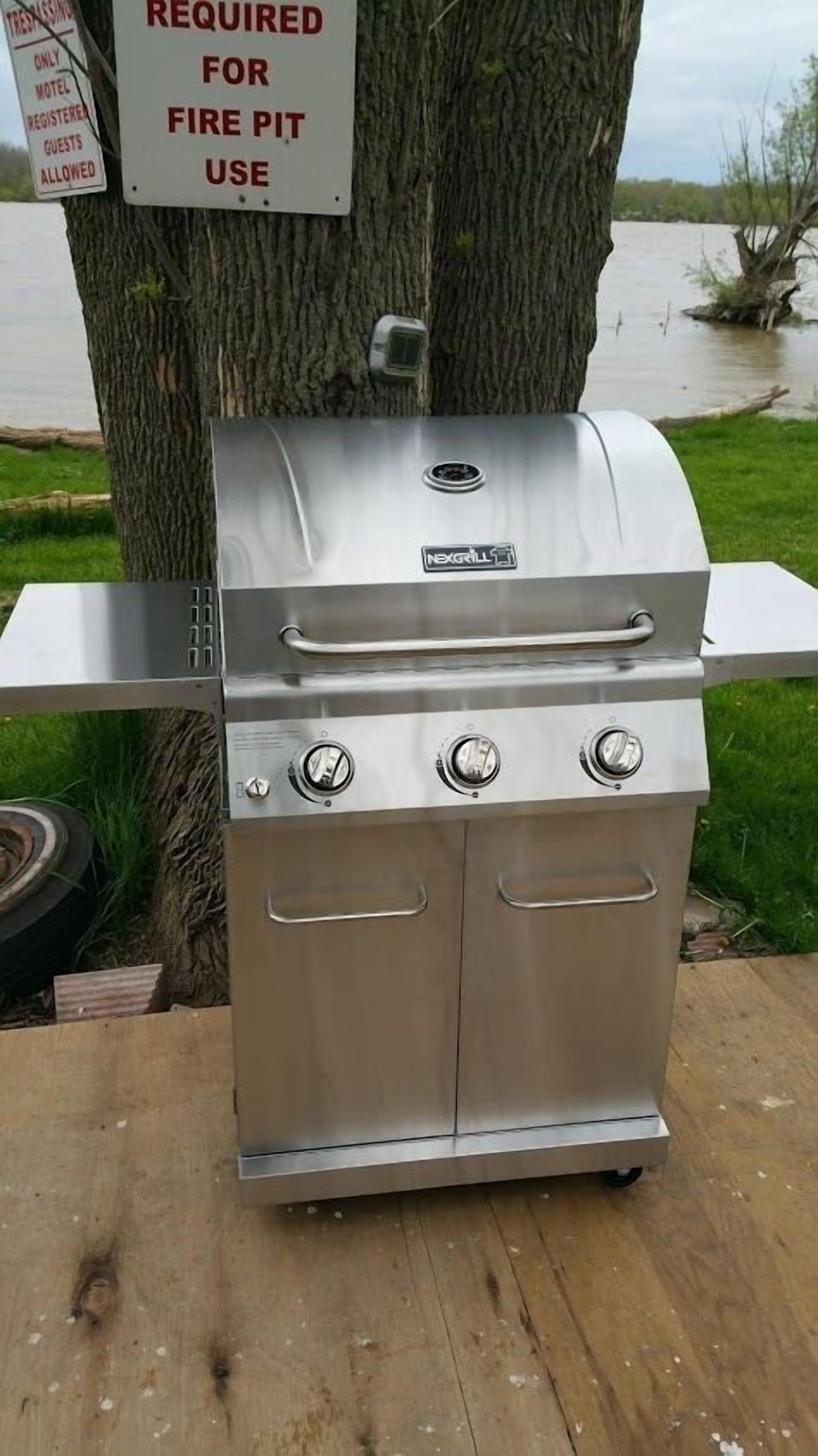 Una parrilla de acero inoxidable con pantalla digital, un quemador lateral y un estante trasero, instalada al aire libre cerca de un árbol y un letrero que indica su uso para fogata.
