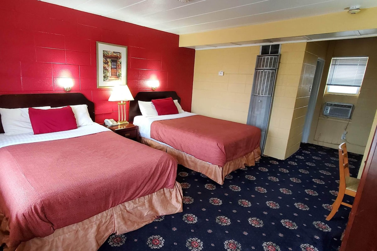 A hotel room with two beds, red walls, a patterned carpet, and a small window with blinds.