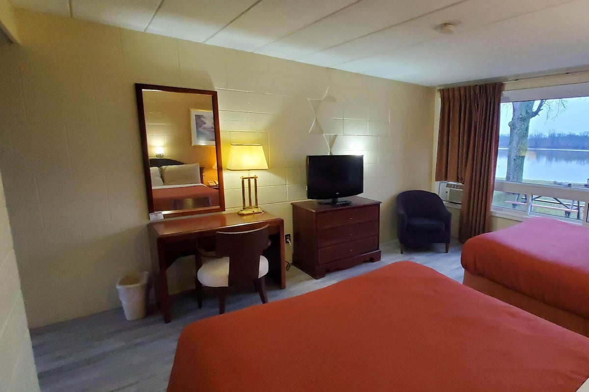A hotel room with two beds, a desk with a chair, a television, a lamp, and a window with a view of a lake.