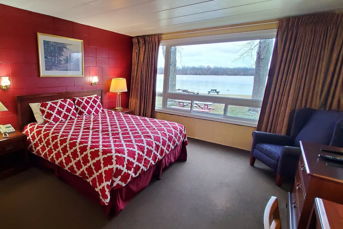 A hotel room with a red brick wall, a large bed with a patterned bedspread, a desk with a chair, a window with a view of a lake and picnic tables, and two bedside lamps.