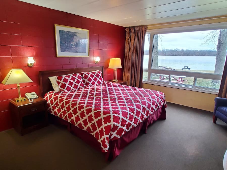 A hotel room with a red brick wall, a large bed with a patterned bedspread, a desk with a chair, a window with a view of a lake and picnic tables, and two bedside lamps.