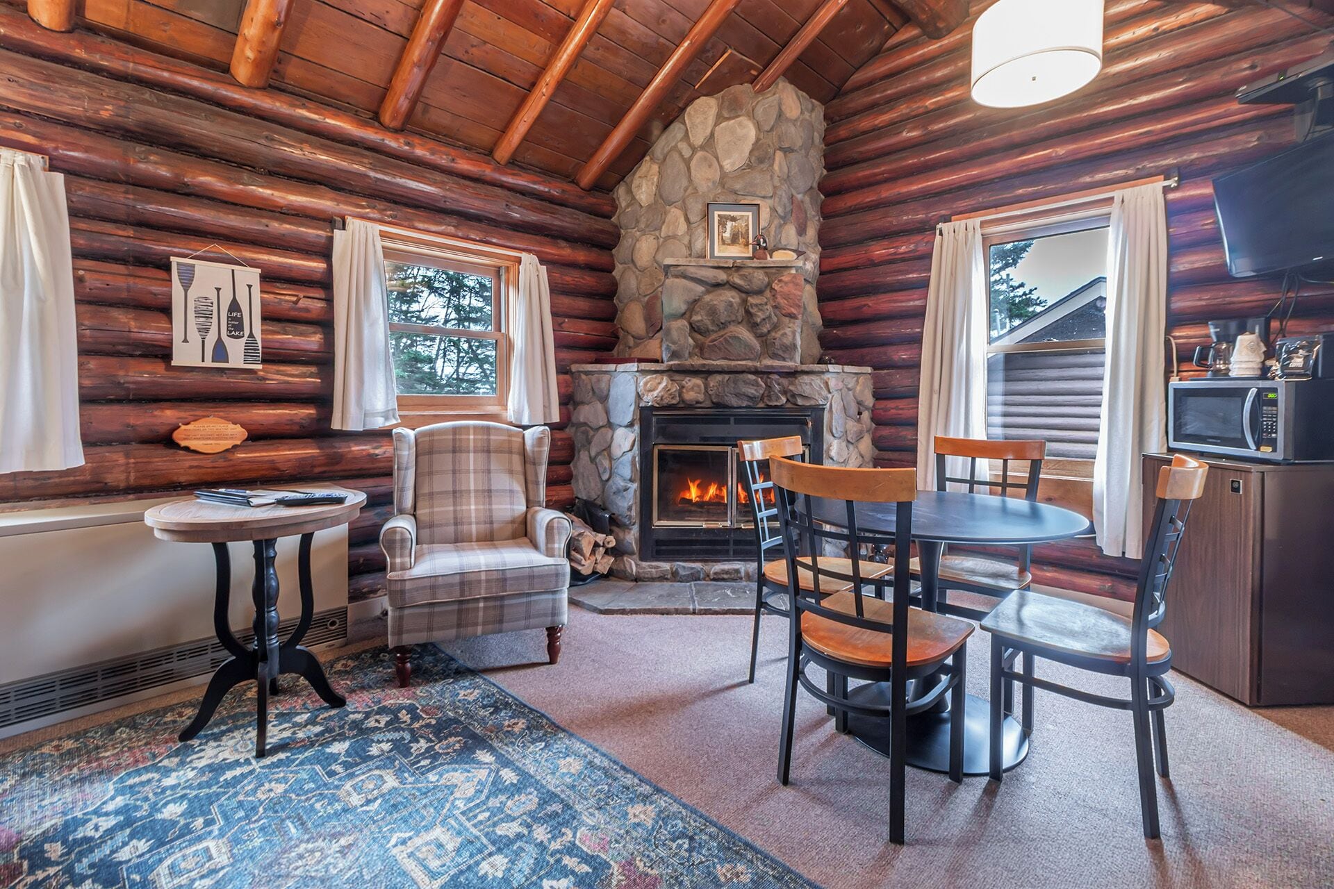 A cozy cabin interior with a stone fireplace, wooden walls, a dining area, and a kitchen with a microwave.