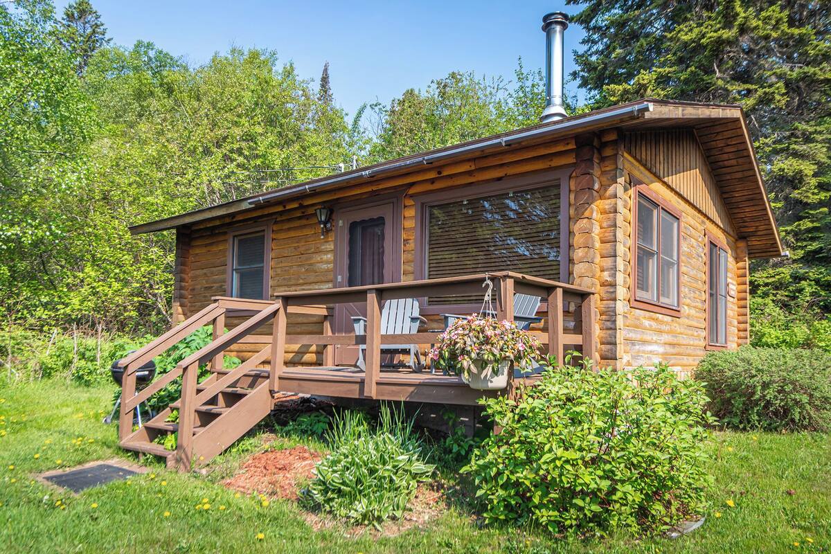 A log cabin with a wooden deck, a staircase, and a hanging flower basket.