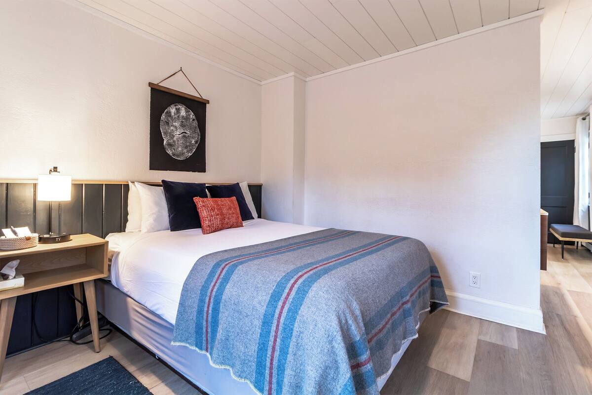 A bedroom with a bed, bedside table, lamp, and a framed artwork on the wall.
