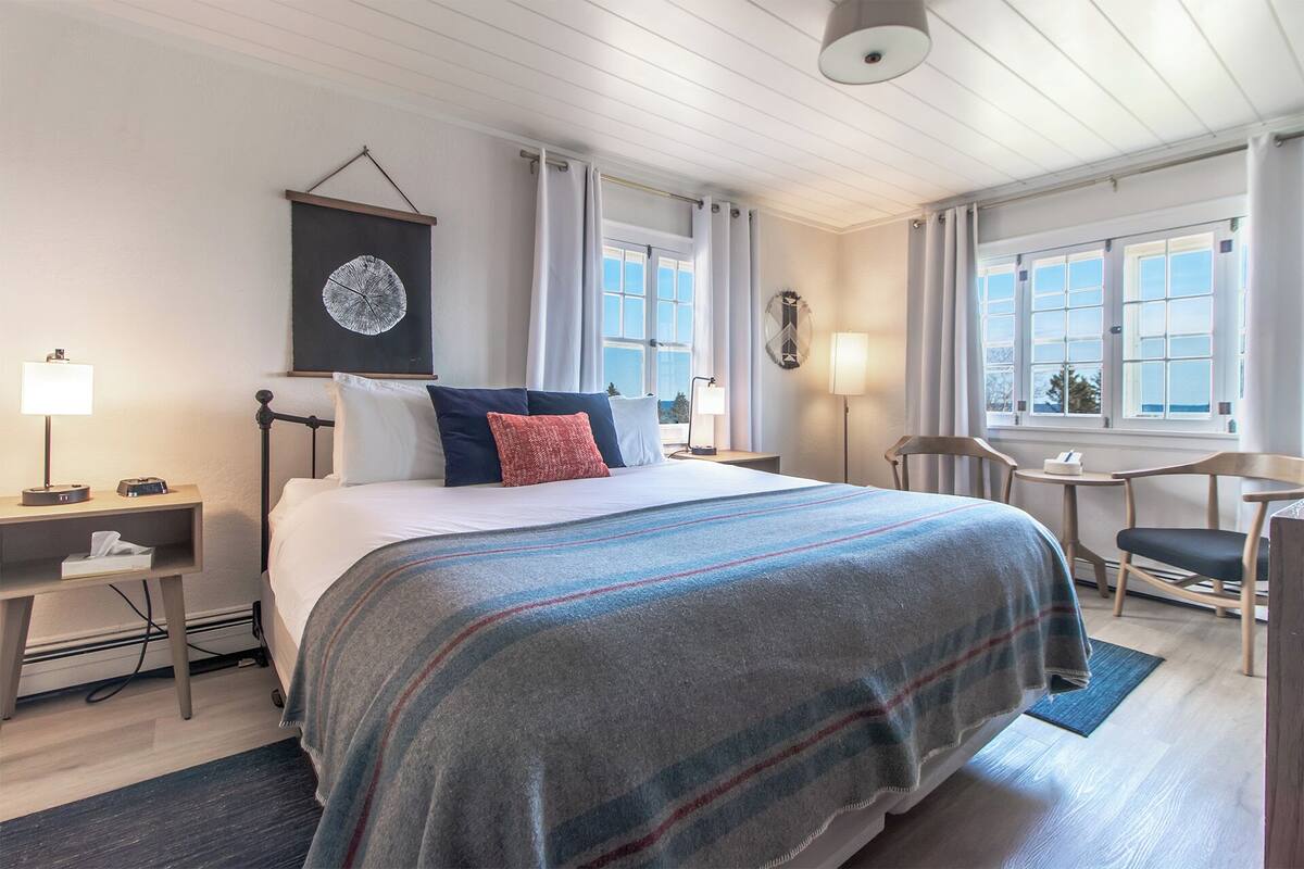A hotel room with a bed, bedside tables, a desk, a chair, and a view of the outside through large windows.