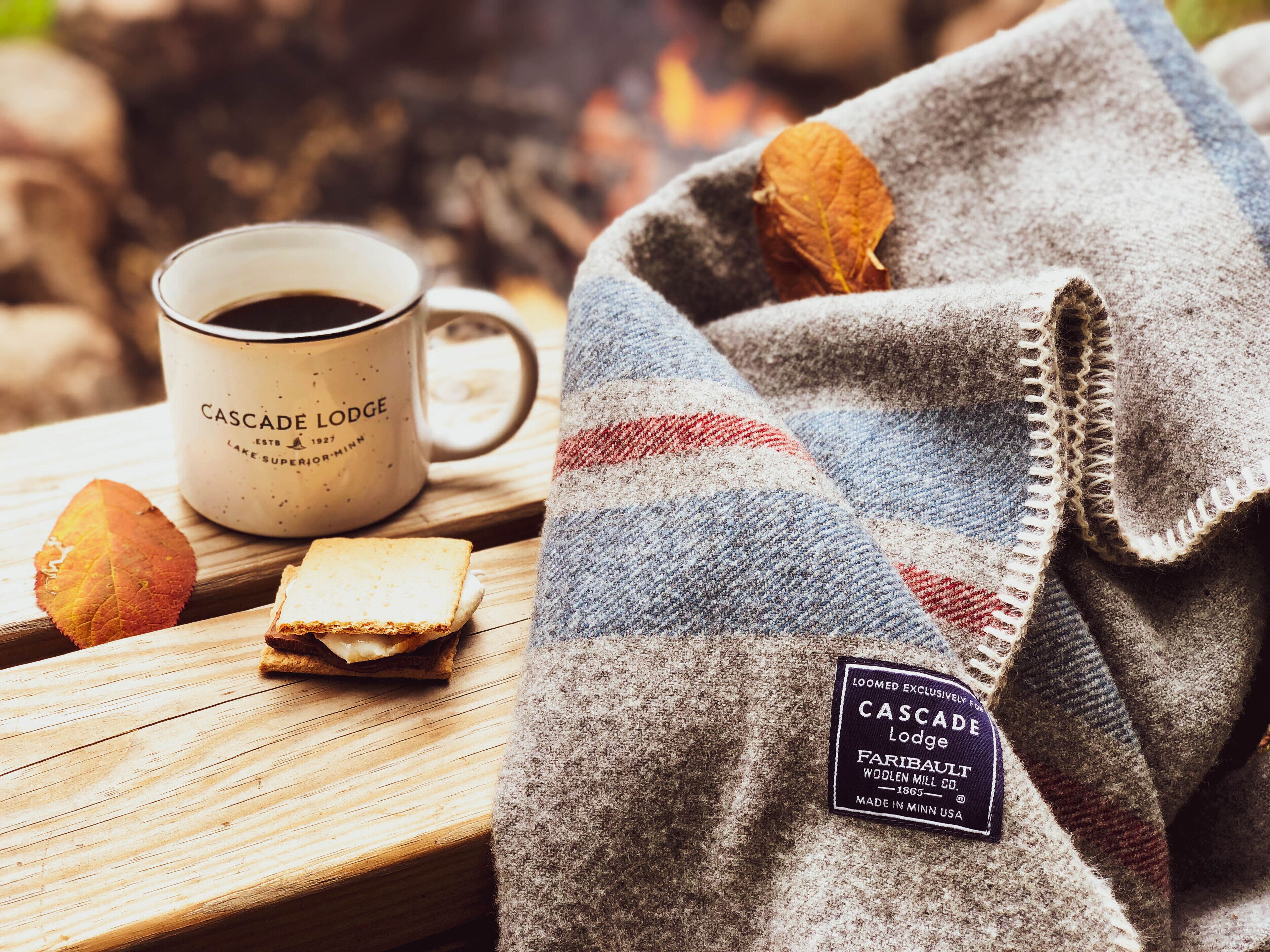 Een houten tafel met een witte mok met de tekst 'Cascade Lodge', een plaid met een label en een stuk brood.
