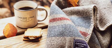 Een houten tafel met een witte mok met de tekst 'Cascade Lodge', een plaid met een label en een stuk brood.