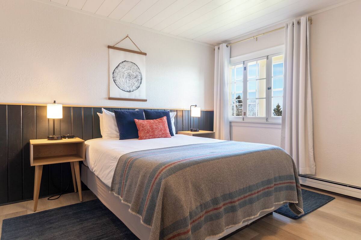 A hotel room with a bed, two bedside tables, a wall-mounted artwork, a window with curtains, and a view of the outside.