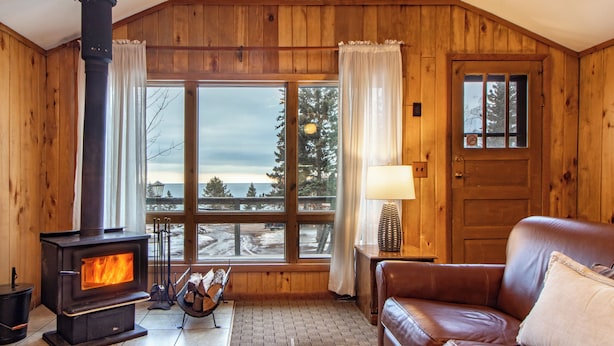 A cozy cabin interior with a wood-burning stove, a leather sofa, and a large window offering a view of the outdoors. - Cascade Lodge (Lutsen)