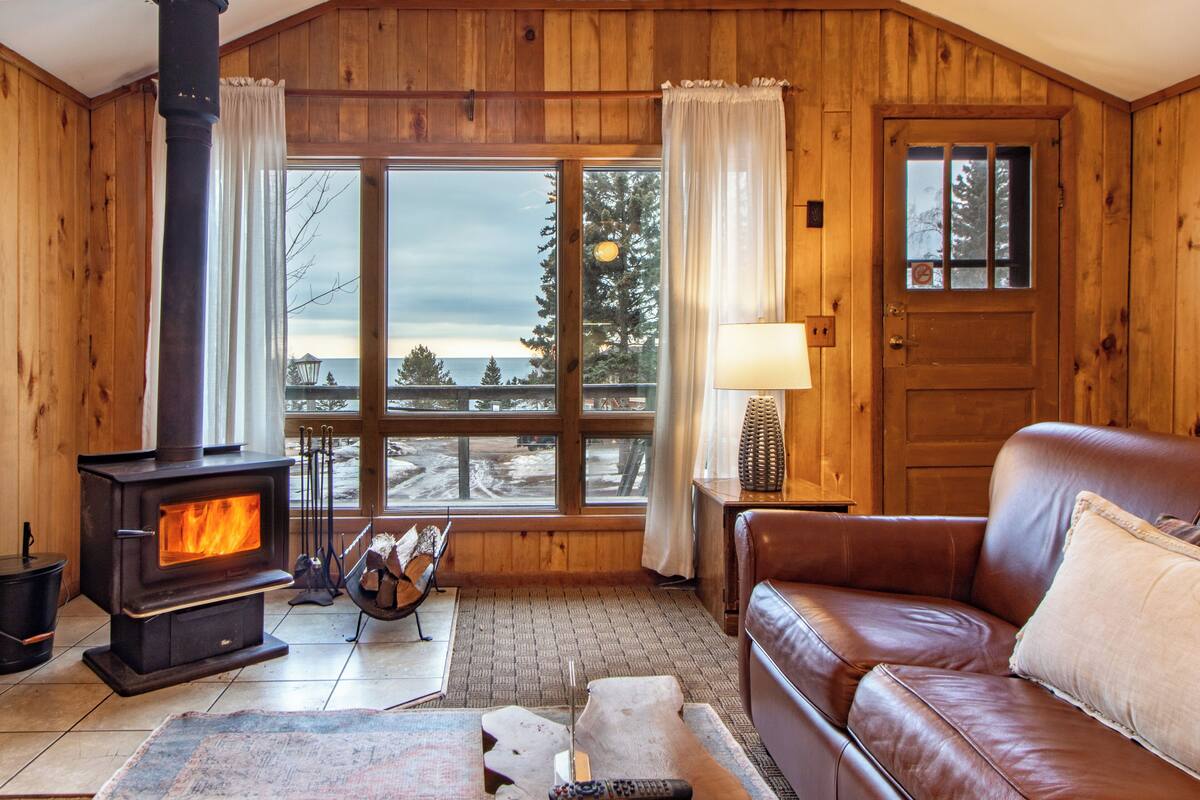 A cozy cabin interior with a wood-burning stove, a leather sofa, and a large window offering a view of the outdoors.