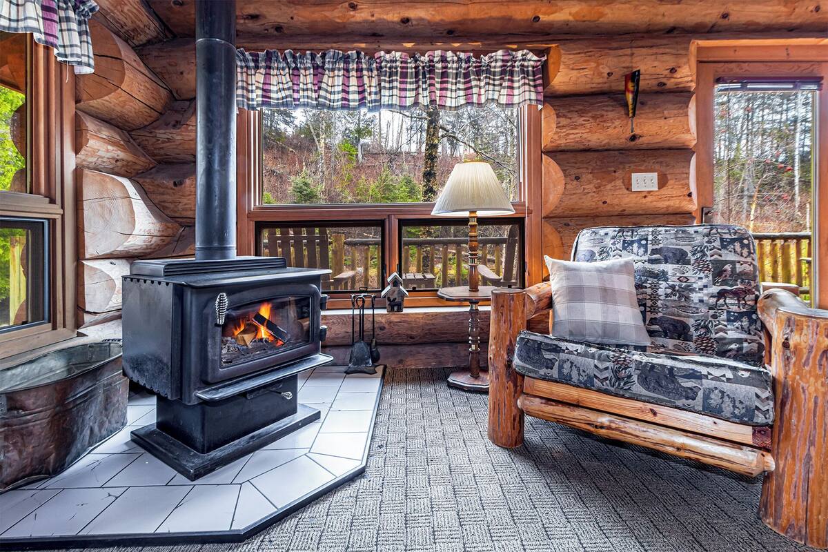 A cozy cabin interior with a wood-burning stove, a cushioned sofa, and a view of the forest through large windows.