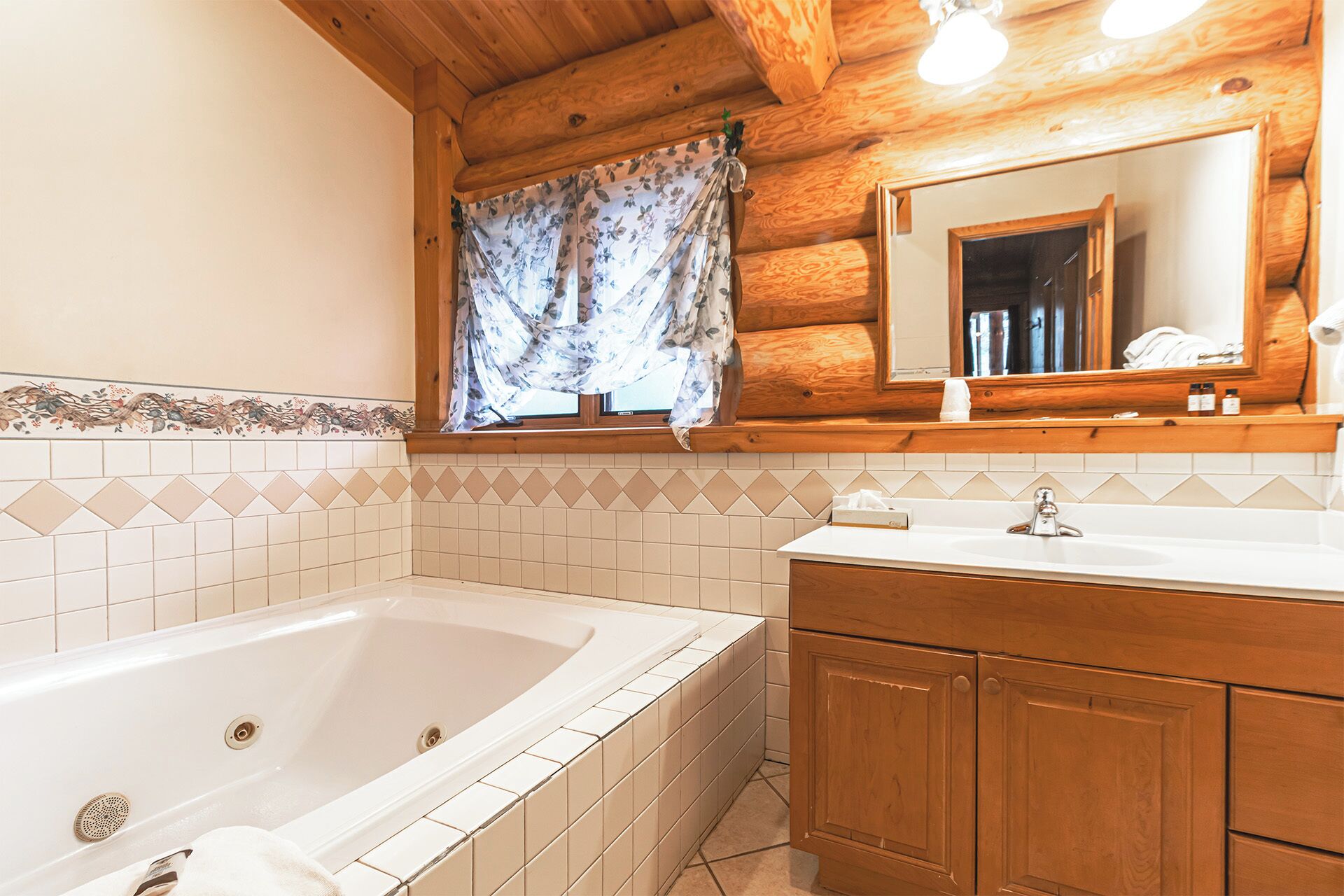 Un baño con techo de madera, una bañera, un lavamanos con espejo y una ventana con cortinas florales.