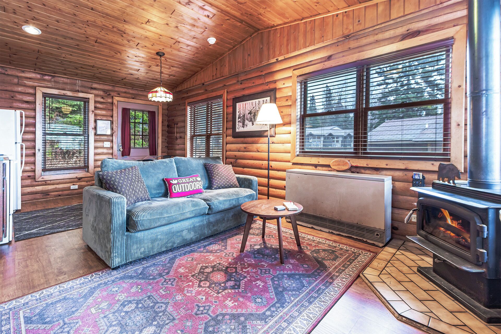 A cozy cabin interior with a sofa, a small table, a fireplace, and a large window with blinds.