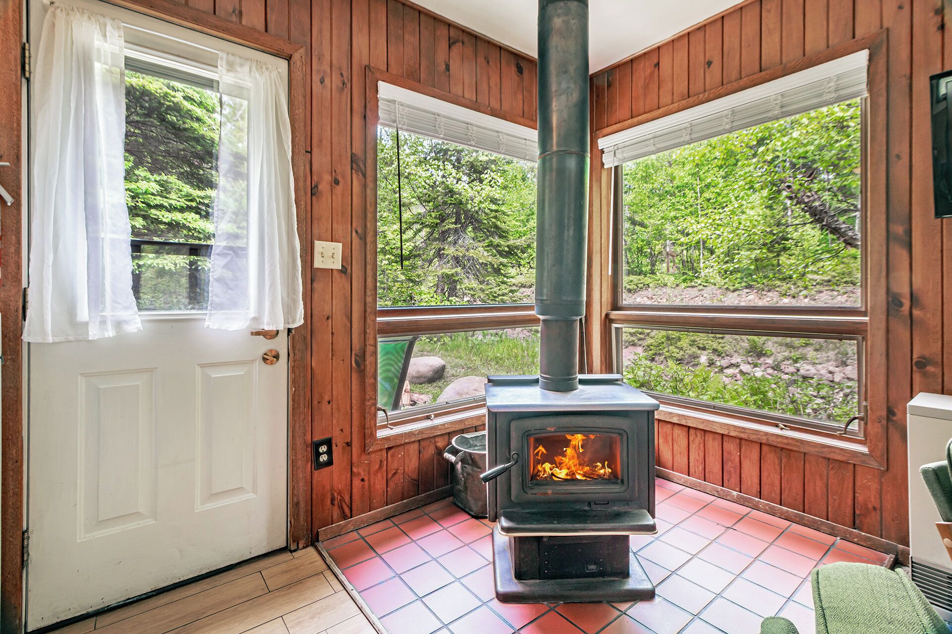 A cozy room with a wood-burning stove, wooden walls, and large windows offering a view of greenery.