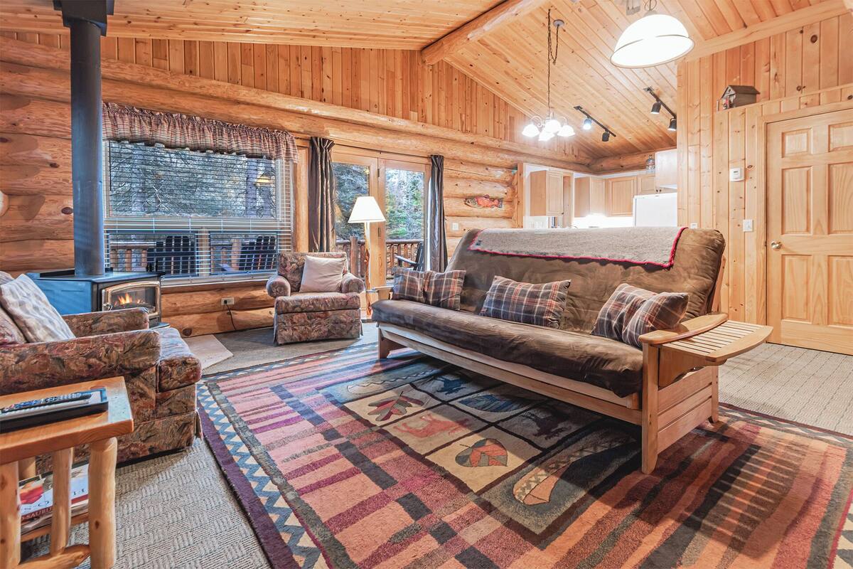 A cozy log cabin interior with a fireplace, a sofa, a chair, and a wooden table.