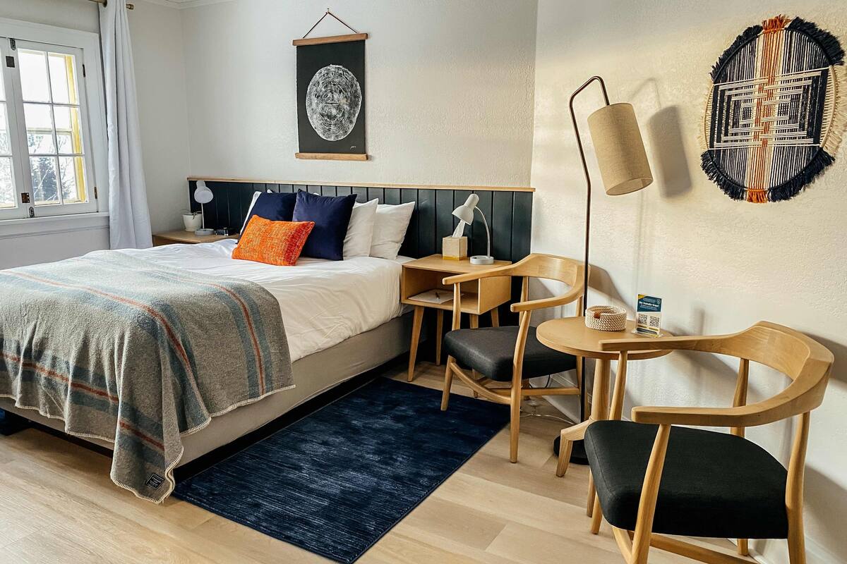 A hotel room with a bed, bedside table, desk, chairs, and a wall-mounted lamp.