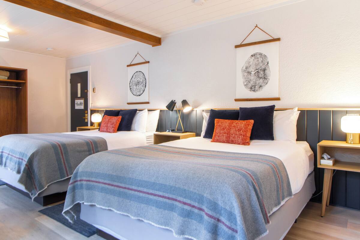 A hotel room with two beds, each with a matching headboard, a bedside table with a lamp, and a framed artwork on the wall.