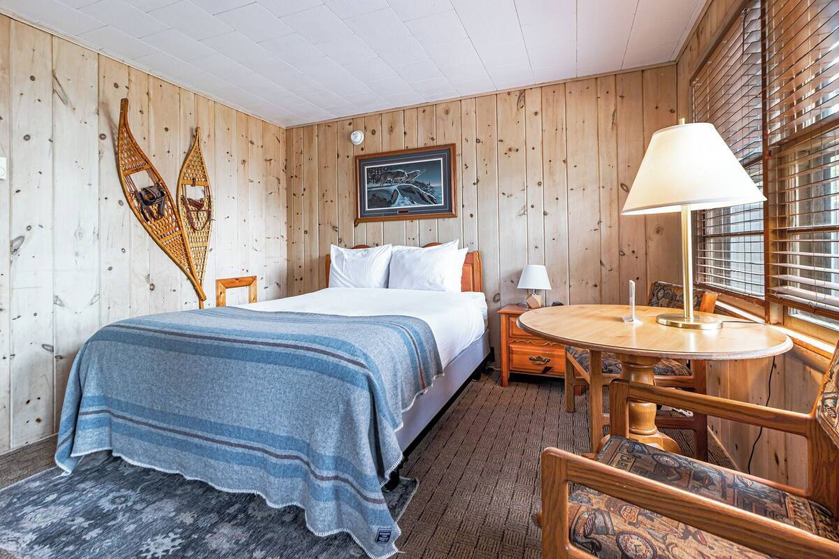 A wooden-paneled room with a bed, a round table with a lamp, and a chair.