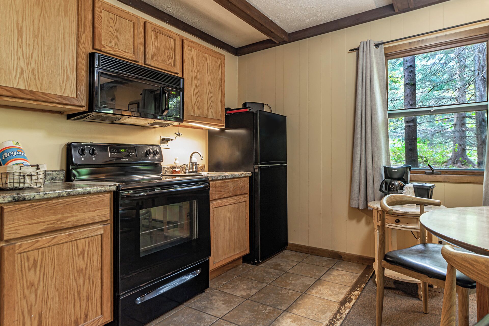 A kitchen with wooden cabinets, a black refrigerator, a stove, and a window with a view of trees.