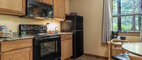 A kitchen with wooden cabinets, a black refrigerator, a stove, and a window with a view of trees.