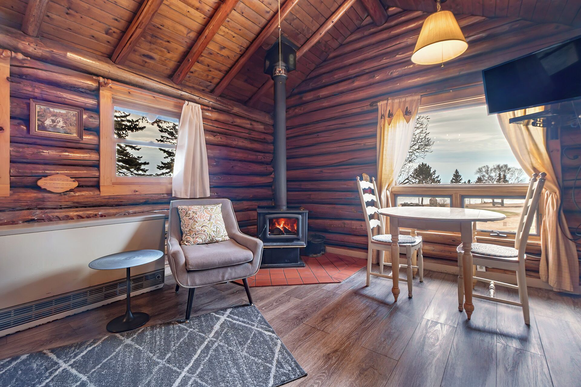 A cozy cabin interior with a fireplace, wooden walls, a dining area, and a view of the outdoors.