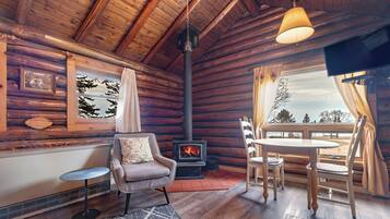 A cozy cabin interior with a fireplace, wooden walls, a dining area, and a view of the outdoors.