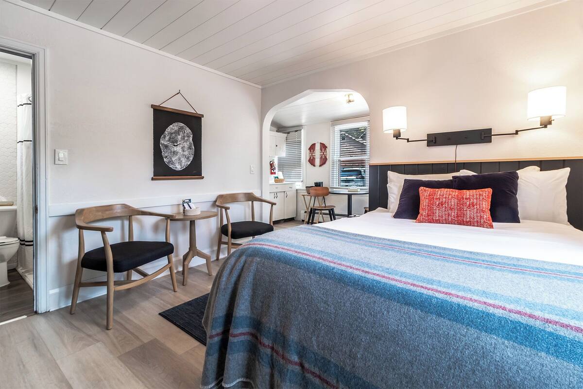 A hotel room with a bed, two chairs, a small table, a wall-mounted artwork, and a window with blinds.