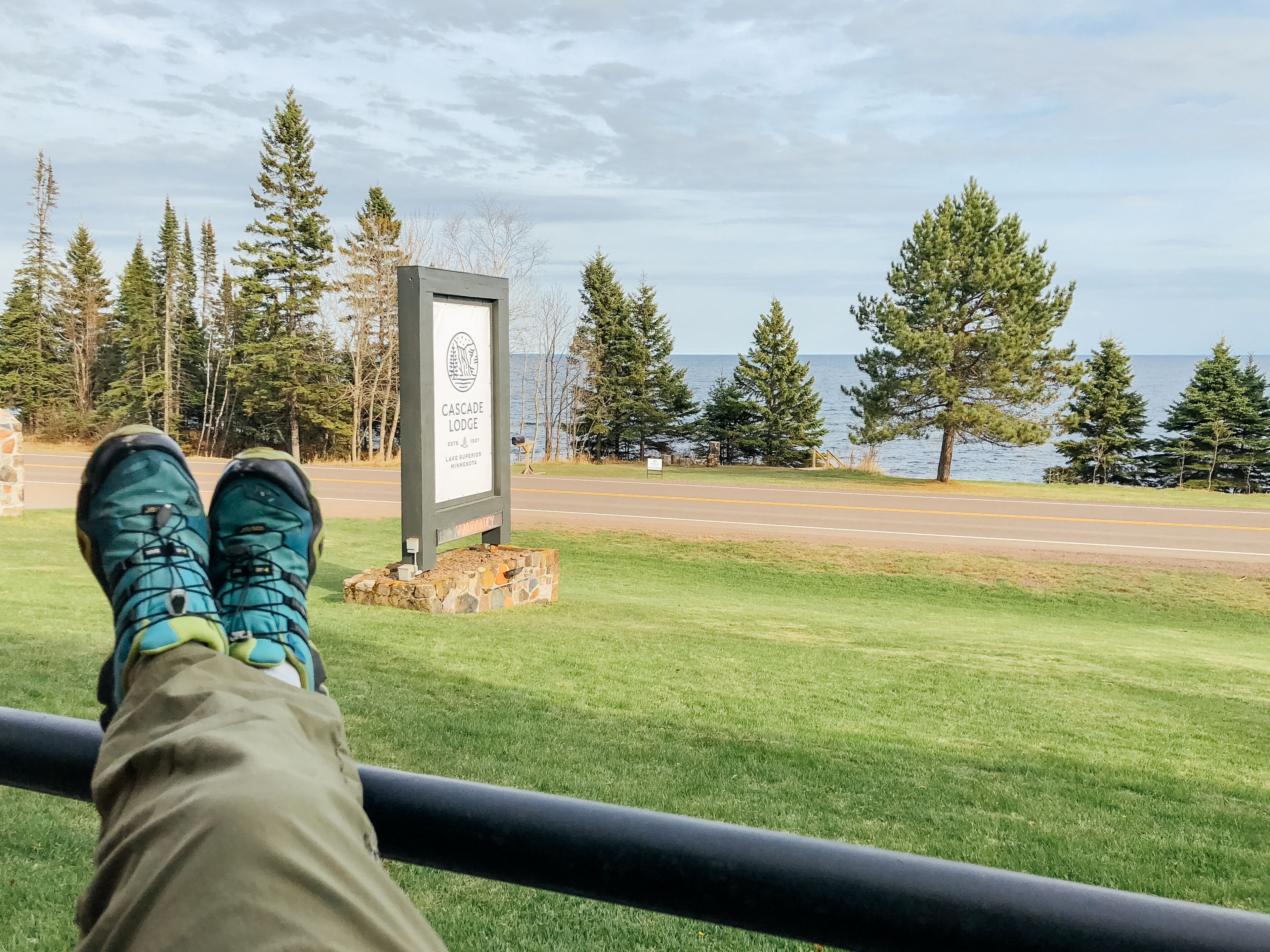 A person relaxing with feet propped up on a railing, overlooking a road with a sign for Cascade Lodge, surrounded by trees and a view of the water.
