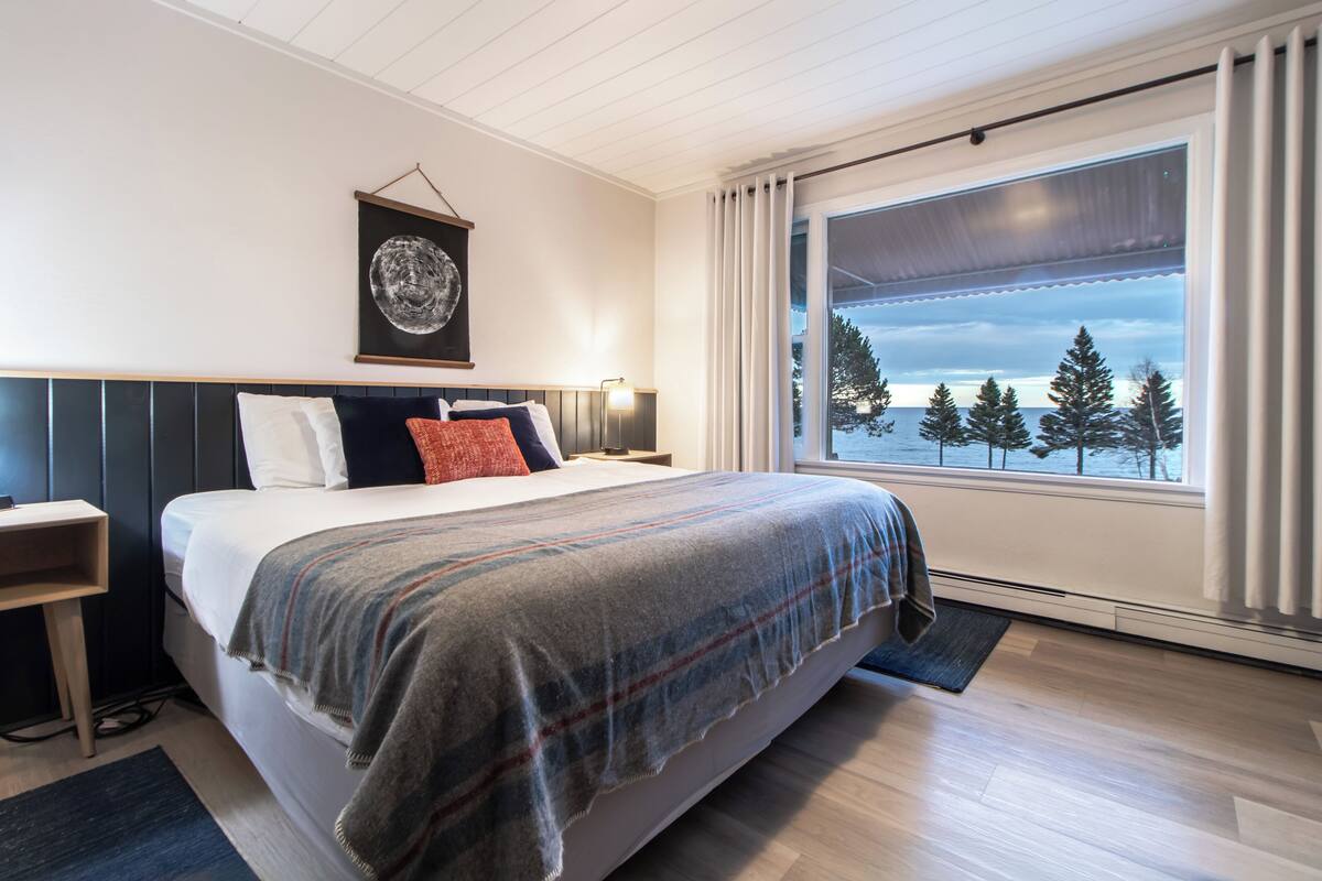A modern bedroom with a large bed, a nightstand, a lamp, and a window with a view of trees and the sky.