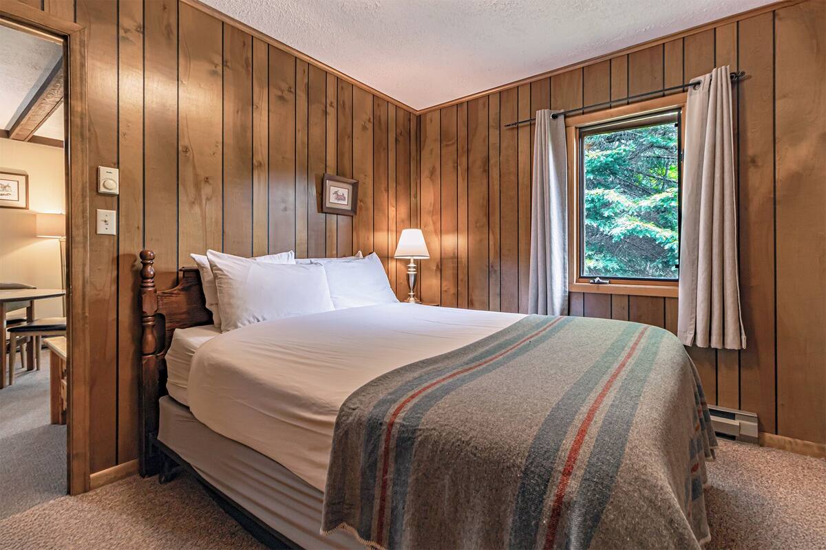 A wooden-paneled bedroom with a bed, a nightstand, a lamp, and a window with curtains.