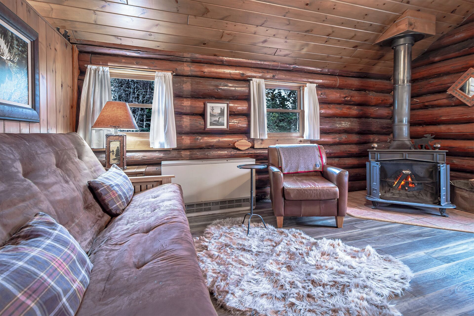 A cozy log cabin interior with a leather sofa, a fur rug, a chair, and a wood-burning stove.