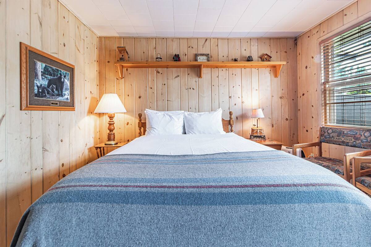 A wooden-paneled room with a bed, two bedside tables with lamps, a chair, and a window with blinds.