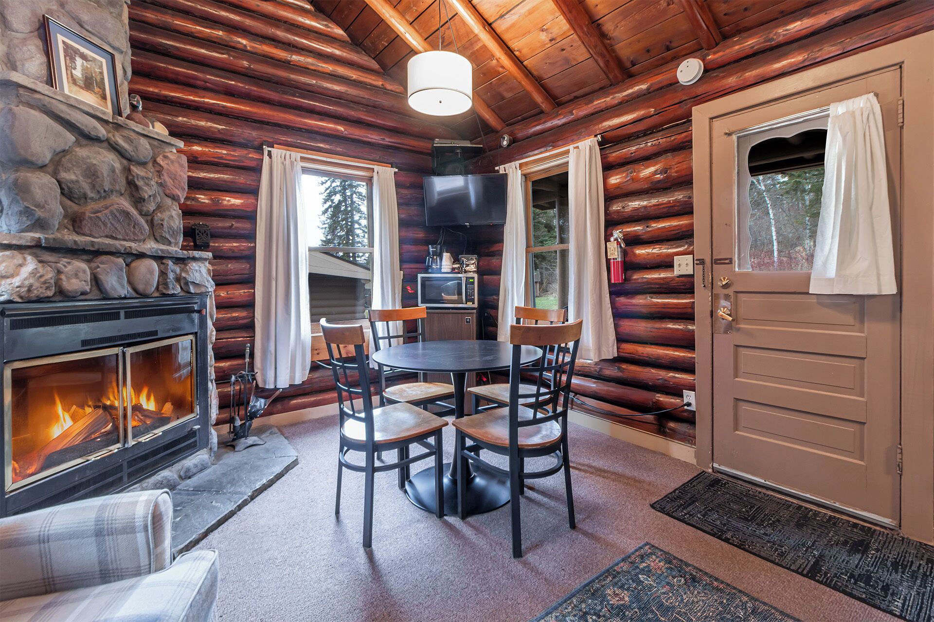 Ein gemütliches Blockhaus-Interieur mit Kamin aus Stein, einem Esstisch mit Stühlen und einer Tür mit Vorhängen.