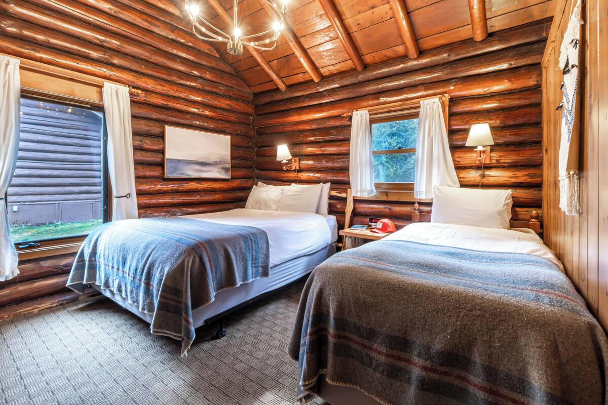 A log cabin room with two beds, wooden walls, a chandelier, and a window with curtains.