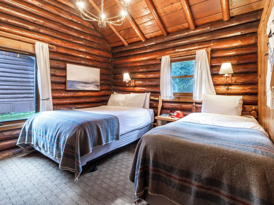 A log cabin room with two beds, wooden walls, a chandelier, and a window with curtains.