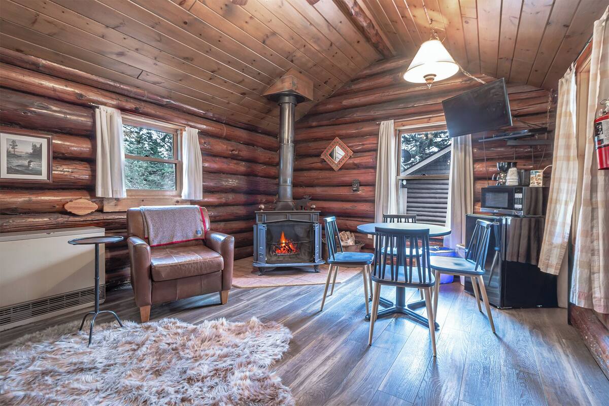 A cozy log cabin interior with a fireplace, leather sofa, dining area, and a kitchen with modern appliances.