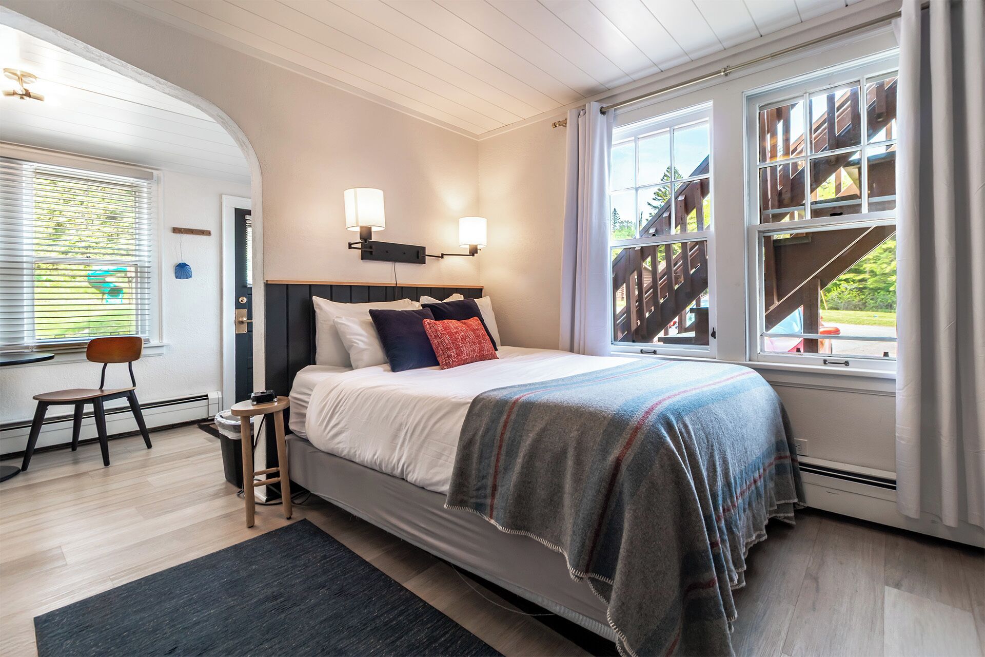 Ein Schlafzimmer mit Bett, Nachttischlampen, einem Sessel, einem kleinen Tisch und einem Fenster mit Blick auf eine Holzterrasse.