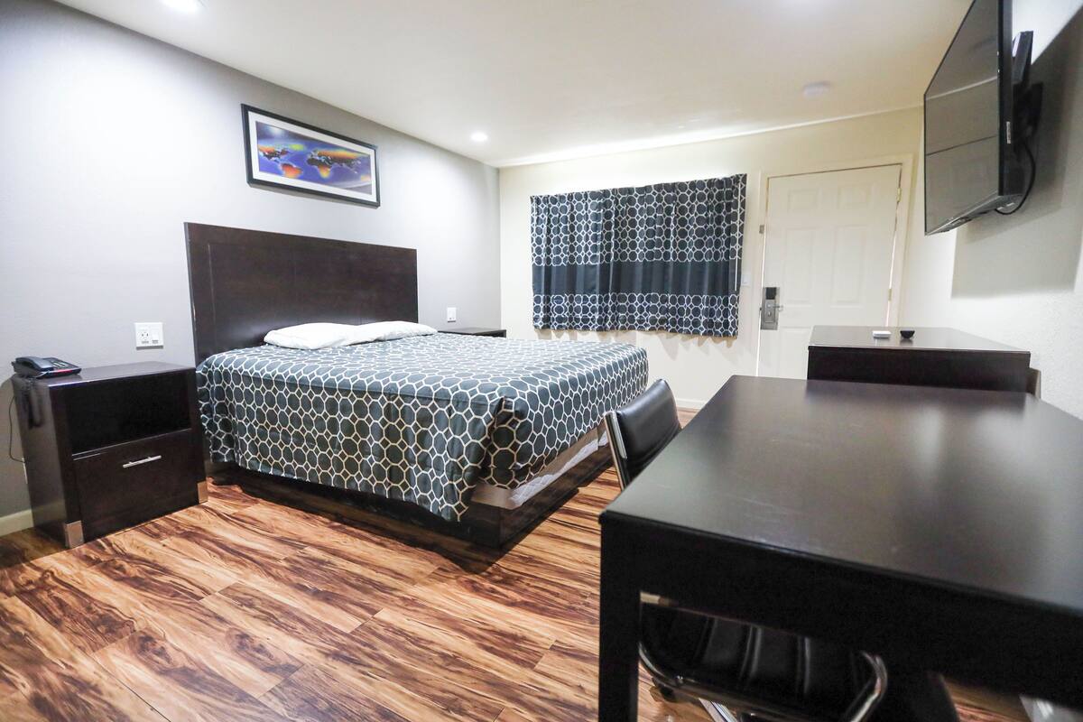 A hotel room with a bed, a desk, a chair, a TV mounted on the wall, and a window with patterned curtains.