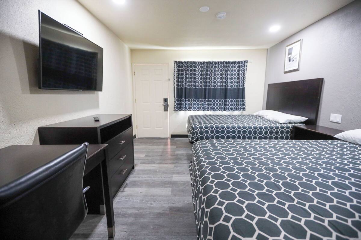 A hotel room with two beds, a desk, a dresser, a flat-screen TV, and a window with patterned curtains.