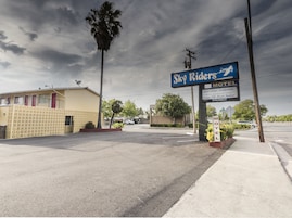 Un motel llamado 'Sky Riders' con un letrero, una palmera y un edificio de fachada amarilla.