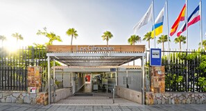 The entrance to Los Girasoles with a stone wall, metal gate, and palm trees. - Relaxia Los Girasoles (San Bartolomé de Tirajana)