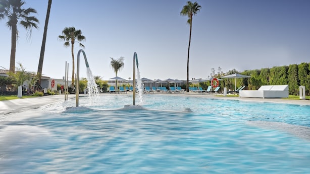 A swimming pool with water fountains, palm trees, lounge chairs, and a white building in the background. - Relaxia Los Girasoles (San Bartolomé de Tirajana)