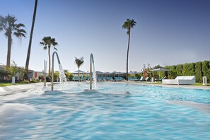 A swimming pool with water fountains, palm trees, lounge chairs, and a white building in the background. - Relaxia Los Girasoles (San Bartolomé de Tirajana)