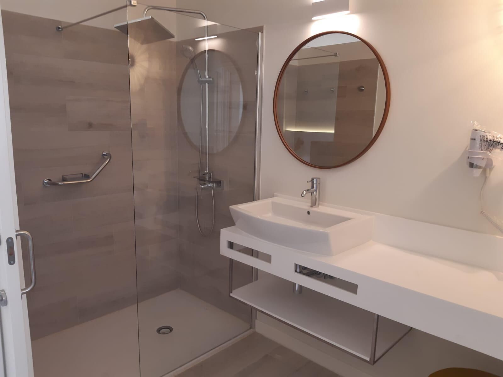 Un baño moderno con ducha envuelta en vidrio, lavamanos blanco y espejo redondo.