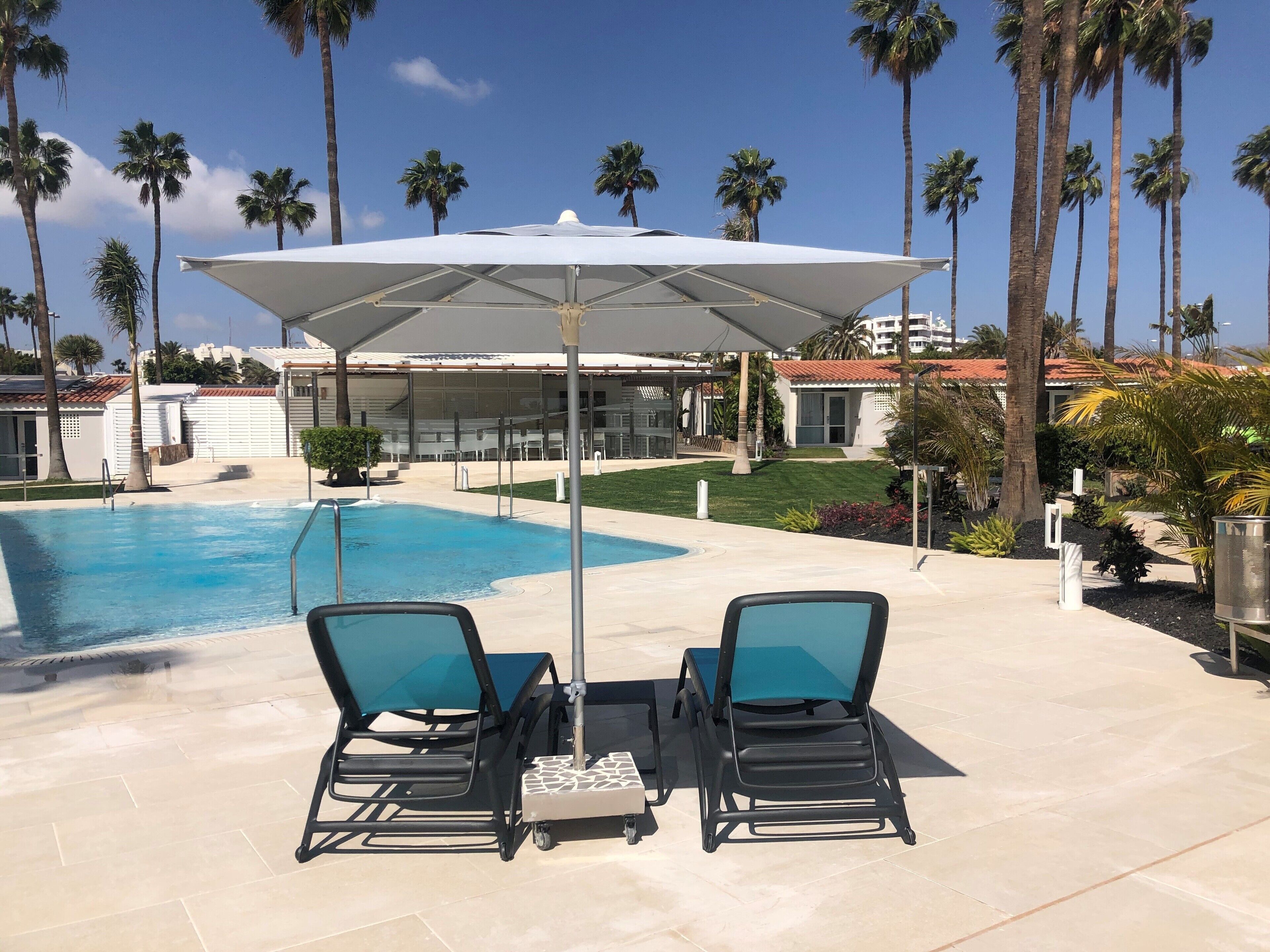 Un espace aménagé au bord de la piscine, avec deux chaises longues et une table placée sous un auvent, le tout entouré de palmiers et d’un bâtiment dont le toit est recouvert de tuiles.