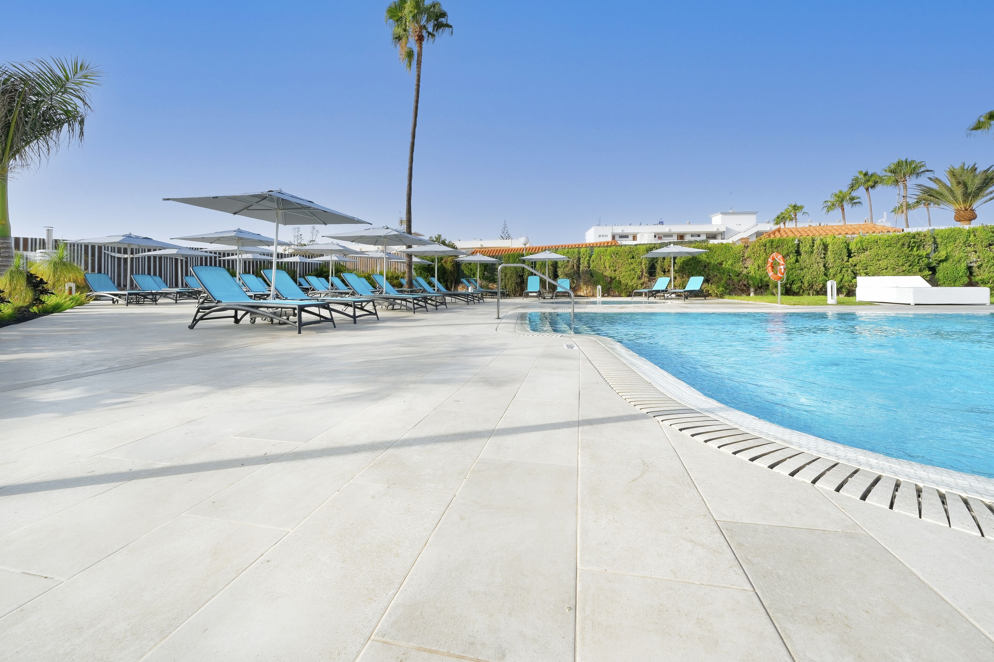A poolside area with lounge chairs and umbrellas, a clear sky, and a building with a tiled roof in the background.